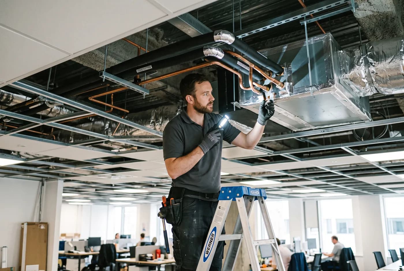 A technician inspecting insulated VRF refrigerant branch pipework above an office ceiling